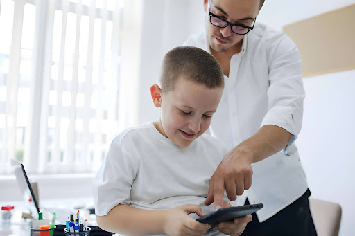 Woman assists a young boy using a tablet at a desk in a bright room, with school supplies nearby.